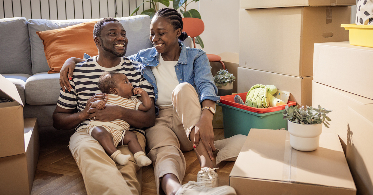 Man and woman sitting on a couch smiling. The man is holding a small baby.