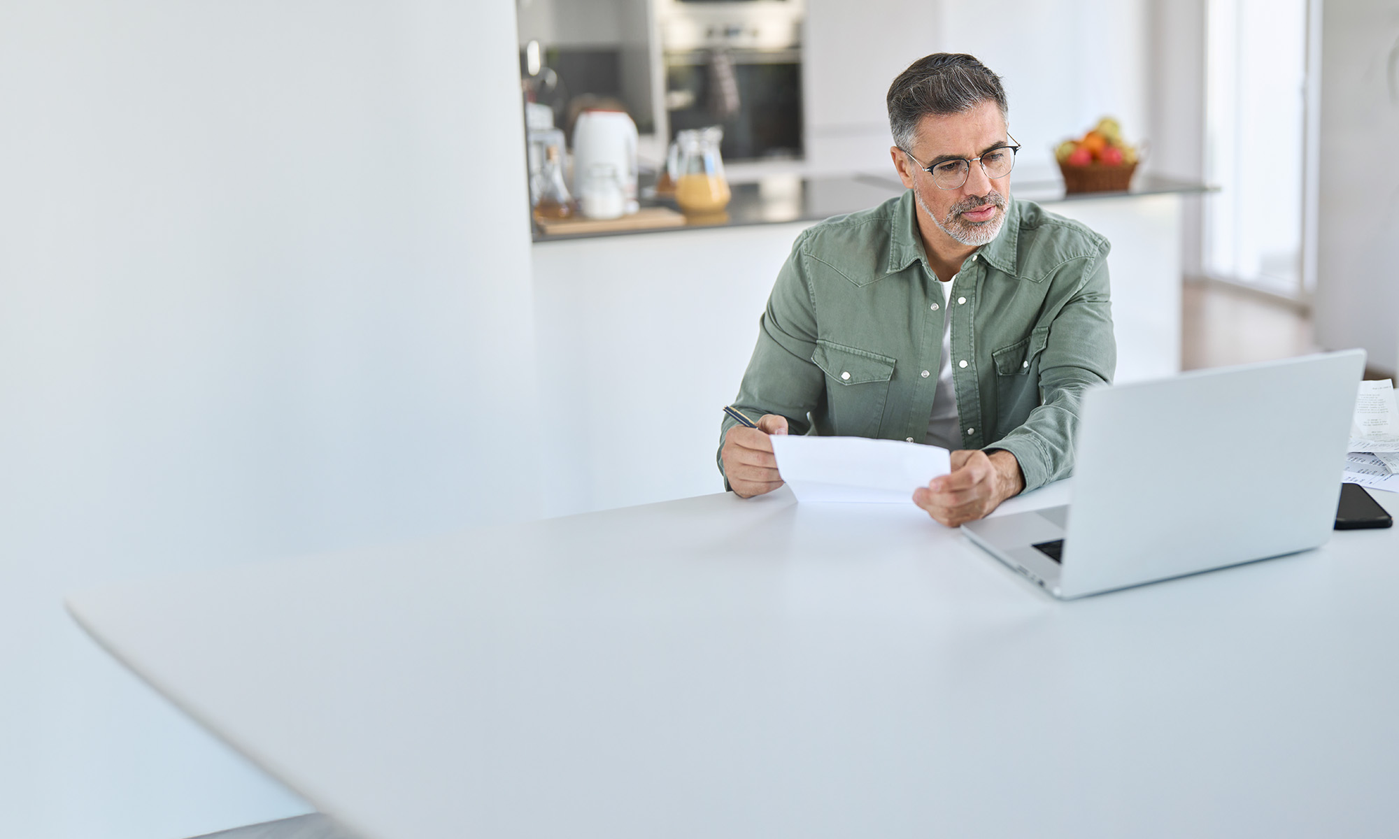 Person sitting at a kitchen table, looking at a laptop and holding paper and pen.