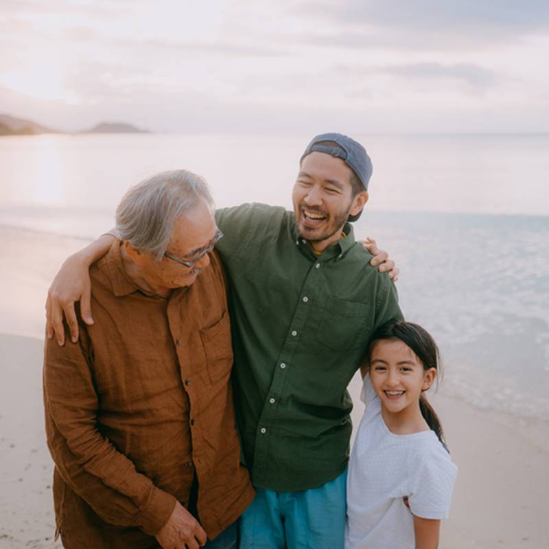 Three people standing together with arms around one another on a rocky shoreline, facing the water.