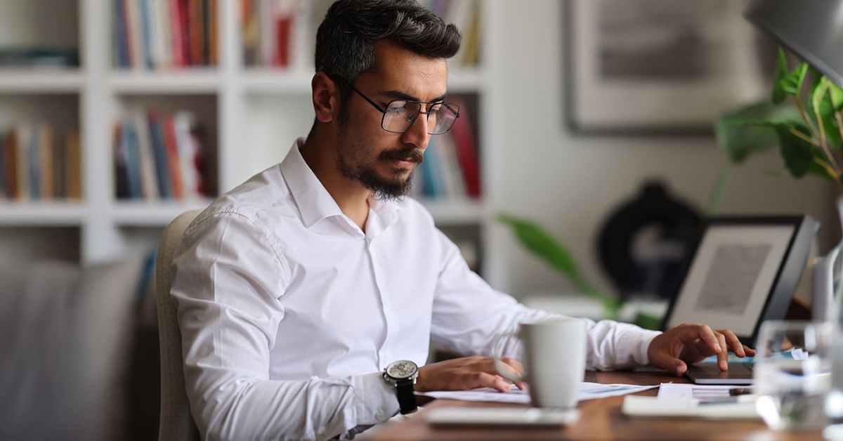 Un jeune homme barbu et portant des lunettes est assis à son bureau à domicile, penché sur une page de renseignements financiers.