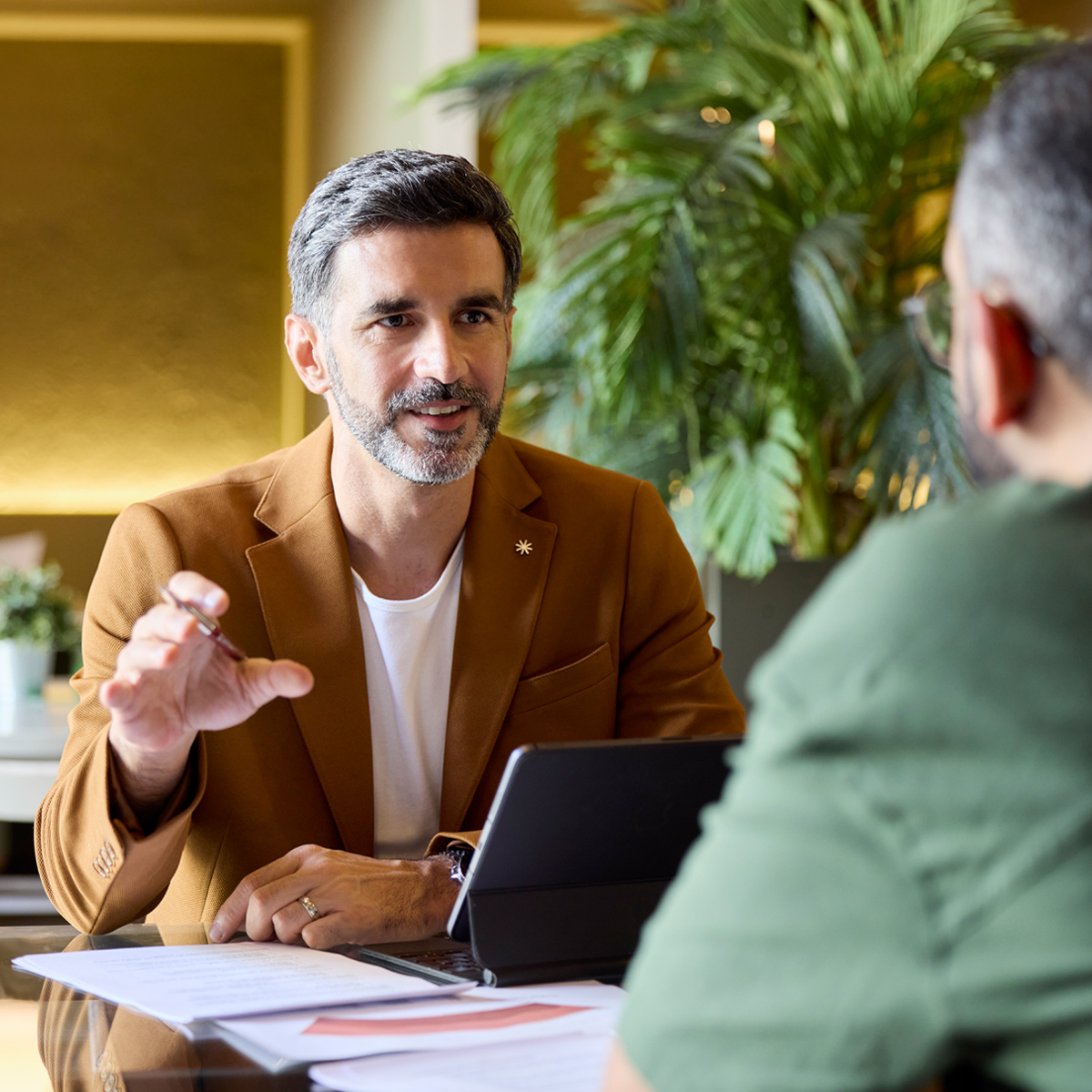 A male financial advisor in a tan blazer smiles and gestures while engaging in a professional conversation with a client.