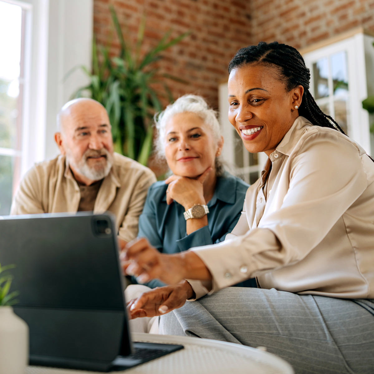 A financial advisor smiles warmly while reviewing information on a laptop with an older couple in a bright, welcoming living room.