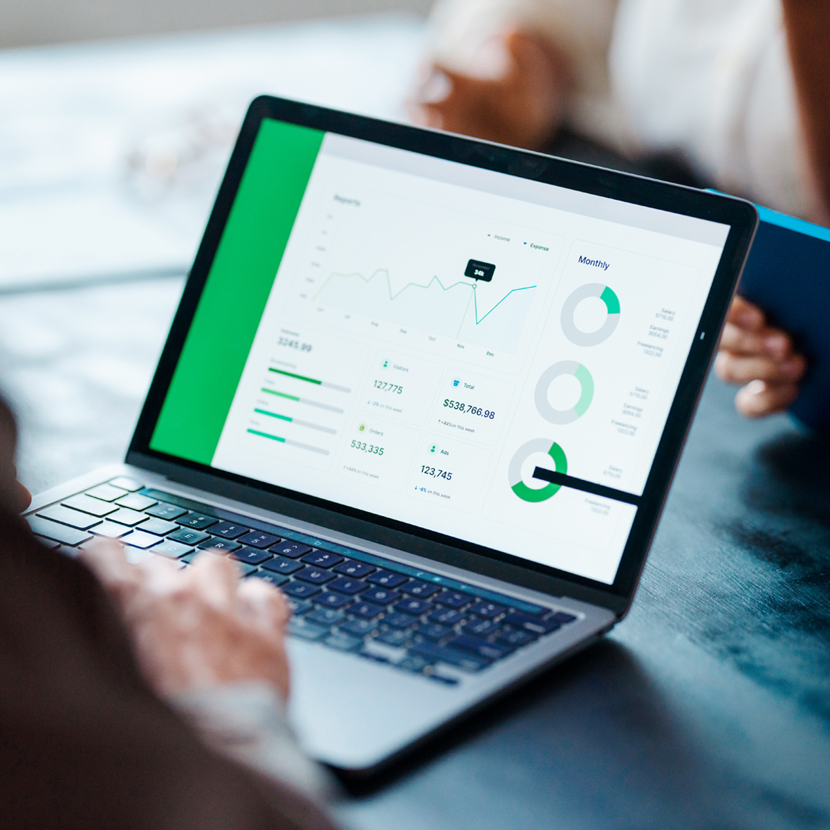 A person's hands rest on a laptop keyboard as a green financial analytics dashboard with charts and performance data is displayed on screen.
