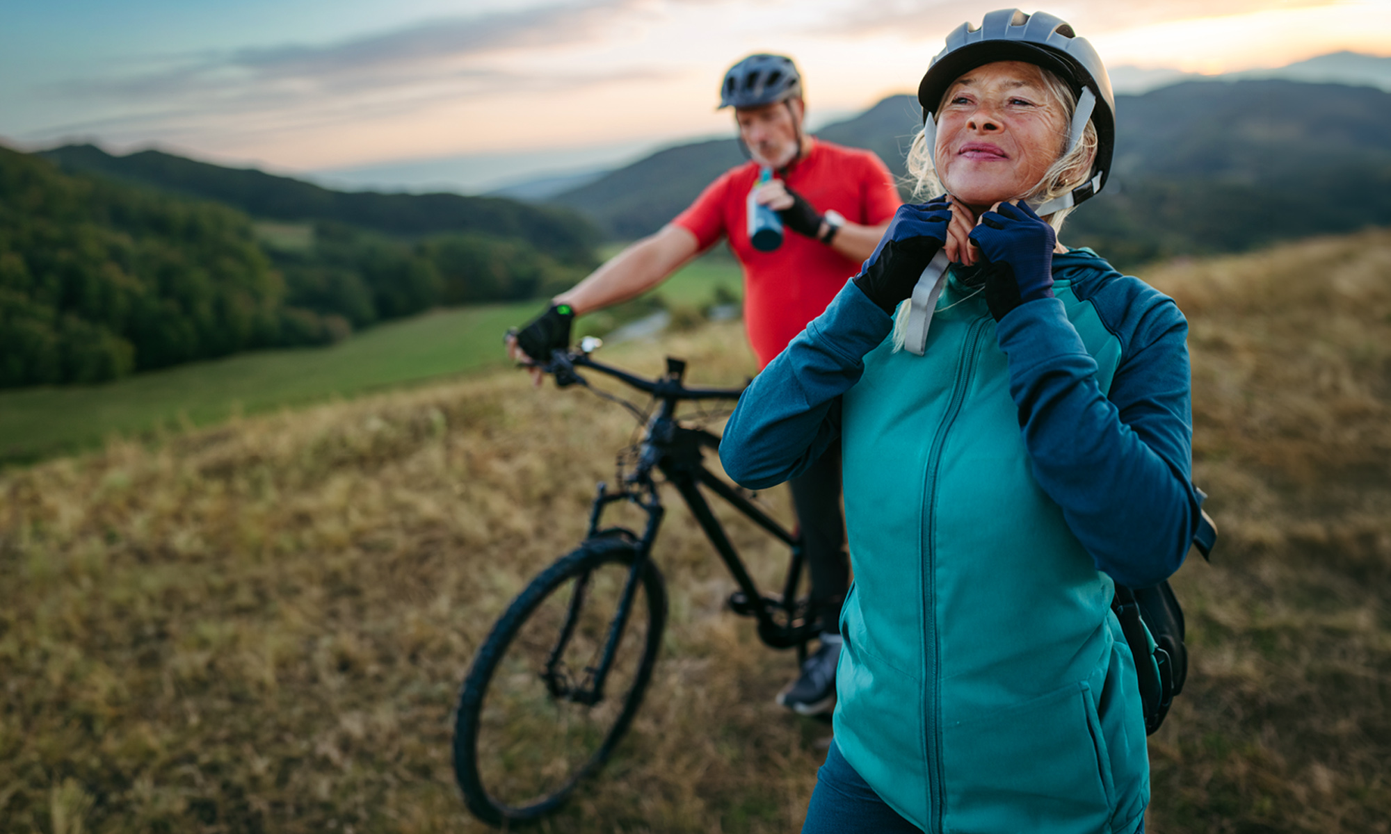 An older woman smiles confidently while adjusting her cycling helmet on a mountain trail, with a man on a mountain bike and a scenic hillside visible behind her.