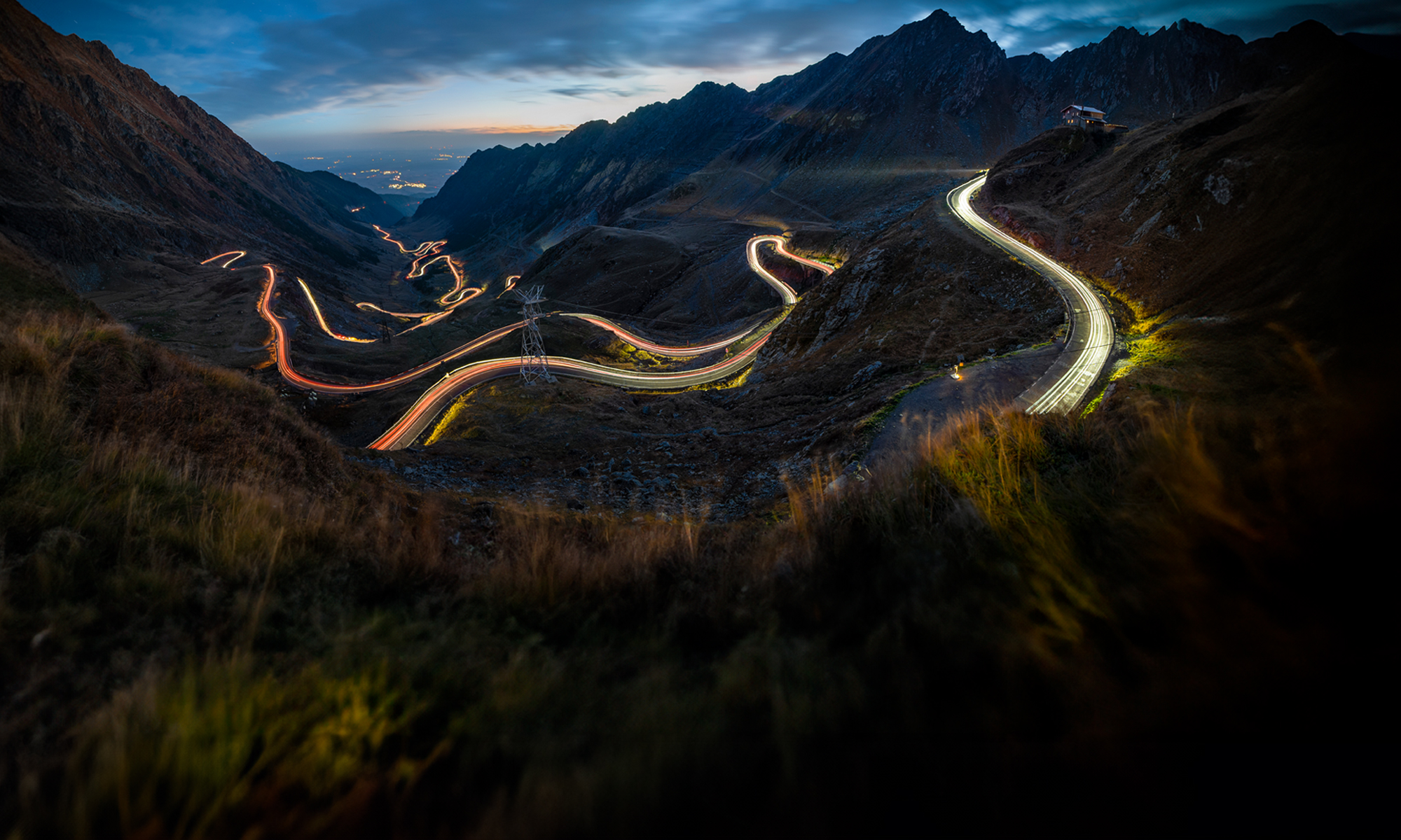 Vue panoramique d’une route de montagne sinueuse au crépuscule, avec des traînées lumineuses laissées par les véhicules en mouvement, formant des motifs fluides sur le paysage accidenté. Les sommets des montagnes se découpent en silhouette sur un ciel assombri, tandis qu’une lueur douce provenant des lumières de la ville est visible à l’horizon lointain.