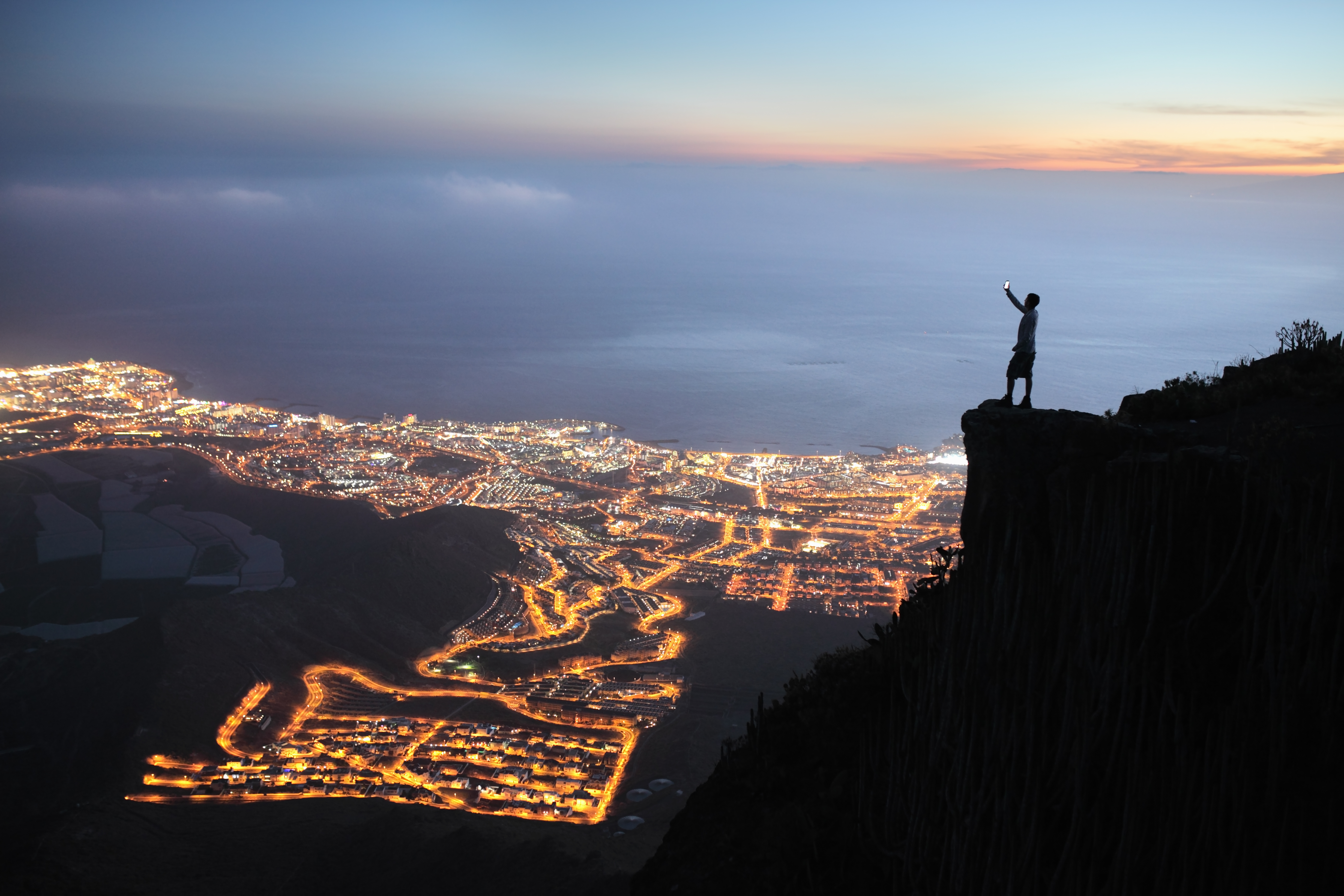 Image of a man standing on the edge of a cliff holding his phone, overlooking a town as the sun sets 