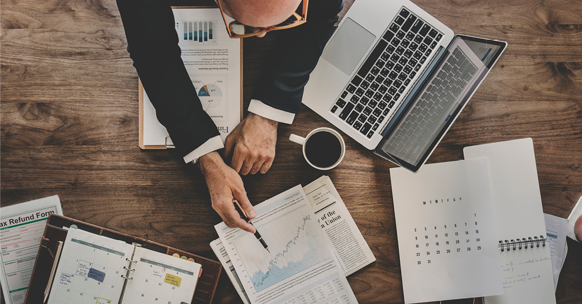 Top-down view of a desk workspace with a person reviewing financial documents and pointing to a chart showing an upward trend. The desk contains one laptop, a cup of coffee, a planner, a calendar, printed reports with graphs, and notes, suggesting financial analysis, business planning, or investment management.