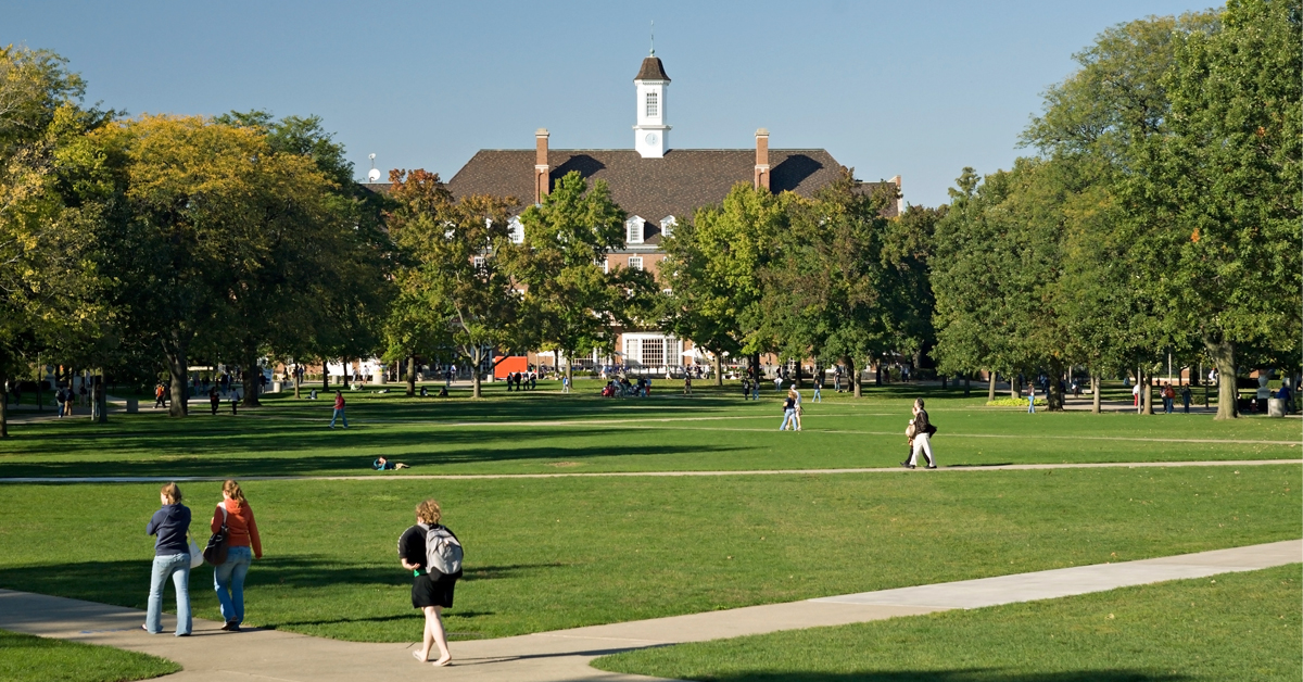 Photo de paysage d’un campus universitaire par une magnifique journée d’automne, avec de l’herbe verte, des arbres, des allées et plusieurs étudiants marchant au premier plan, et un bâtiment administratif ou de salles de classe à l’arrière-plan.