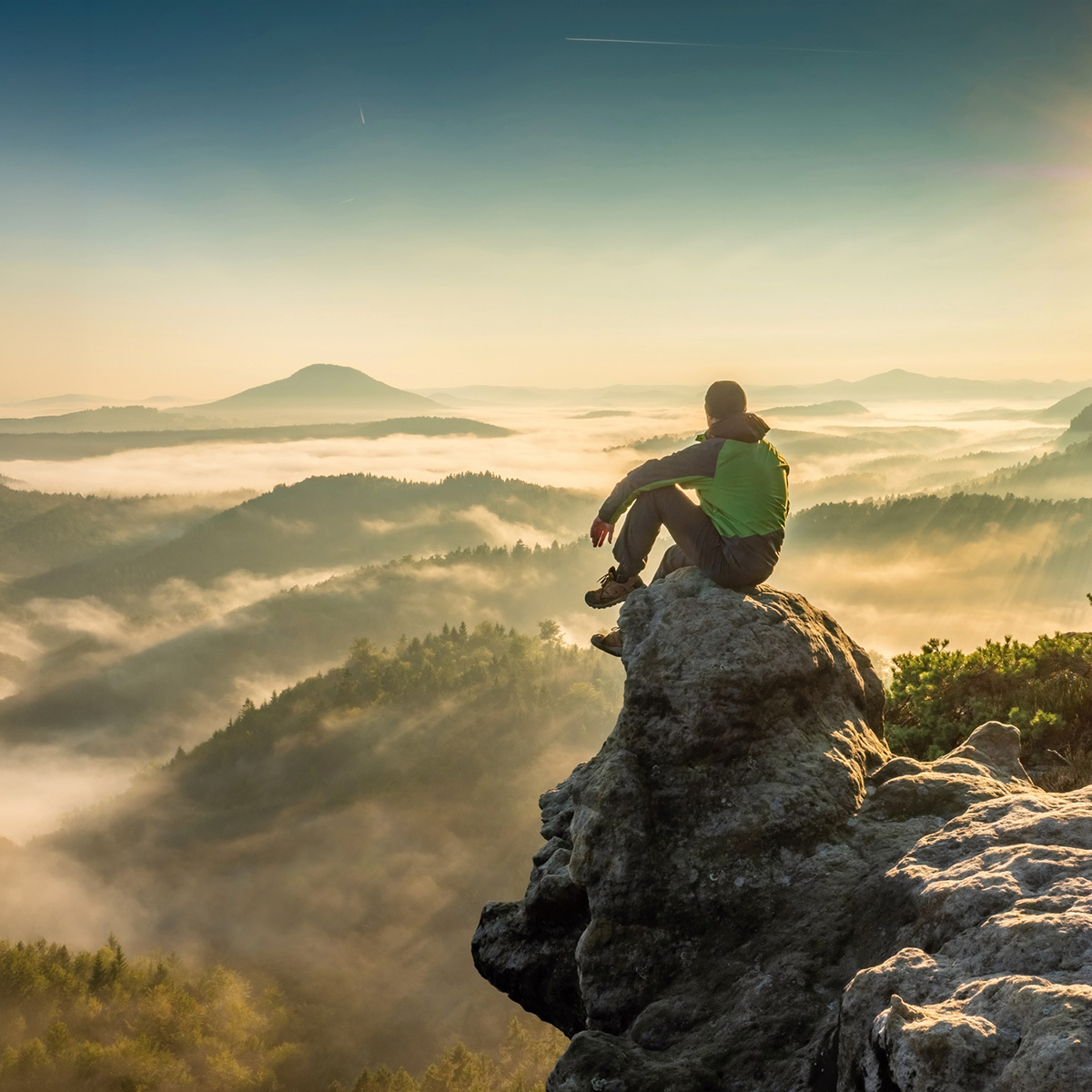 Square-format photo of a man meditating on a hazy summit after hiking, atop a craggy peak with backpack resting nearby.